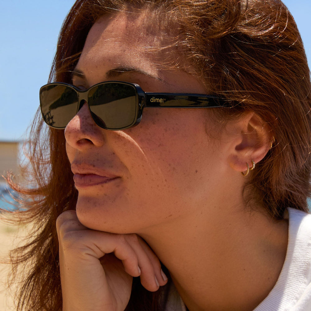 on model - female at the beach wearing dime optics featuring the agoura rectangular sunglasses with a black frame and grey lenses angled view