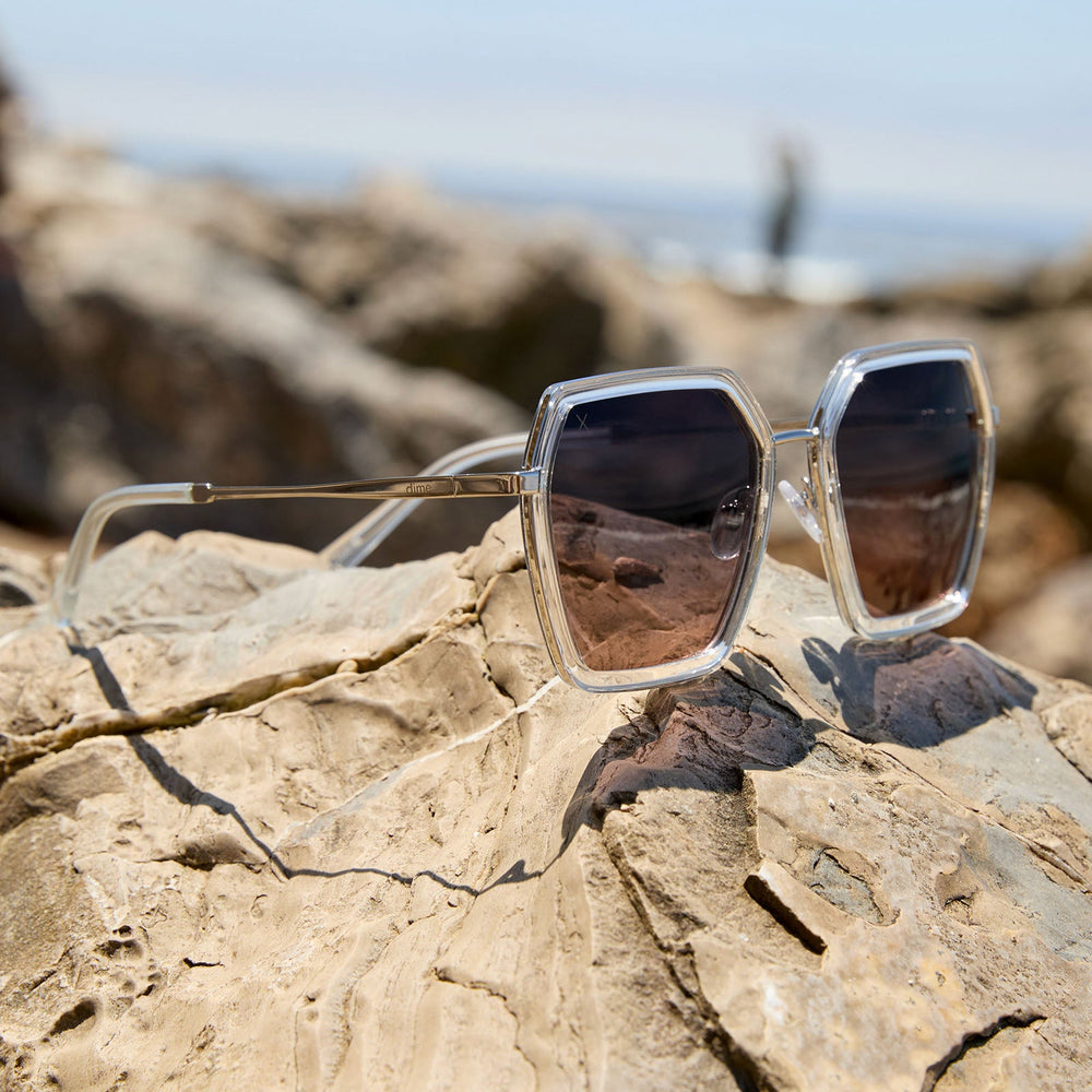 flat lay at the beach of dime optics featuring the alameda square sunglasses with a silver and crystal clear frame and orchid gradient lenses