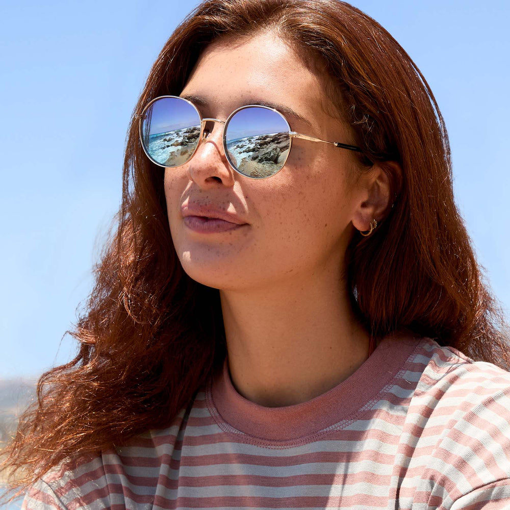 on model - female wearing dime optics featuring the silverlake round sunglasses with a gold frame and wisteria mirror light blue purple lenses angled view