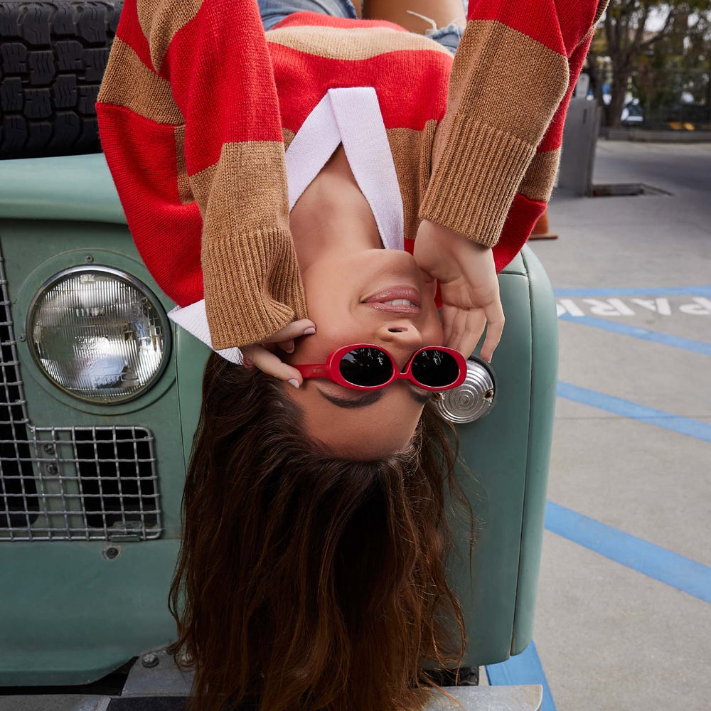 on model - female upside down on a car wearing dime optics westwood round sunglasses with a ruby red frame and grey polarized lenses front view